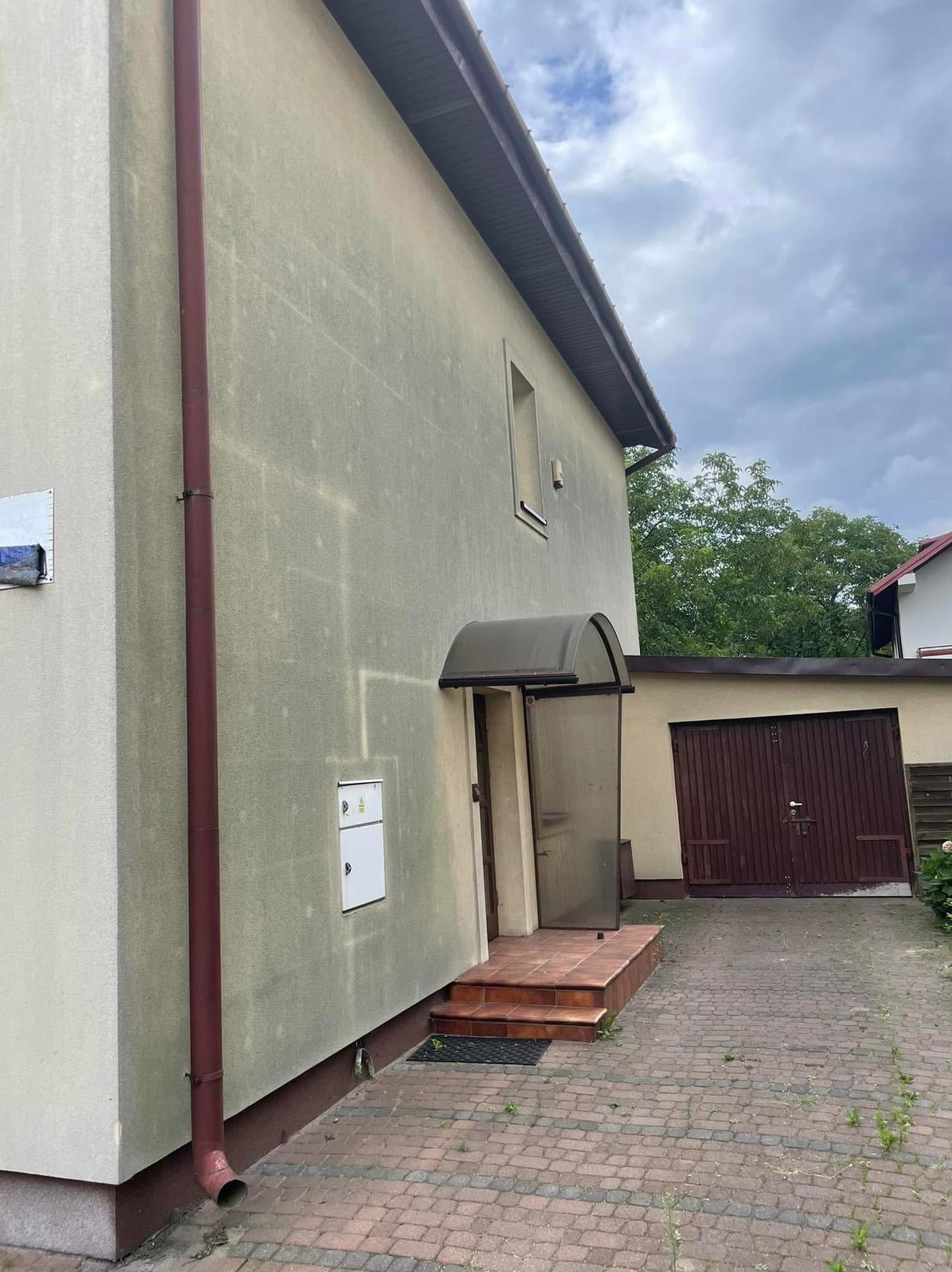 Brudna elewacja domu z widocznymi śladami zabrudzeń i zacieków, brązowa rynna, kostka brukowa przed garażem. Widok z dołu, pochmurne niebo.