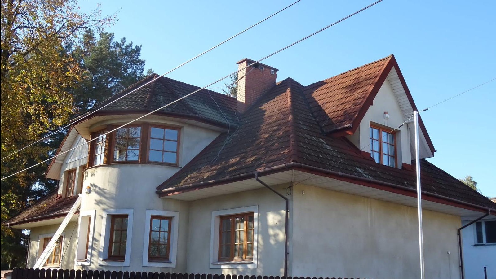 Dwupiętrowy dom z czerwonym dachem, widoczne zabrudzenia na dachu i elewacji. Przystawiona drabina sugeruje przygotowania do prac na wysokości.