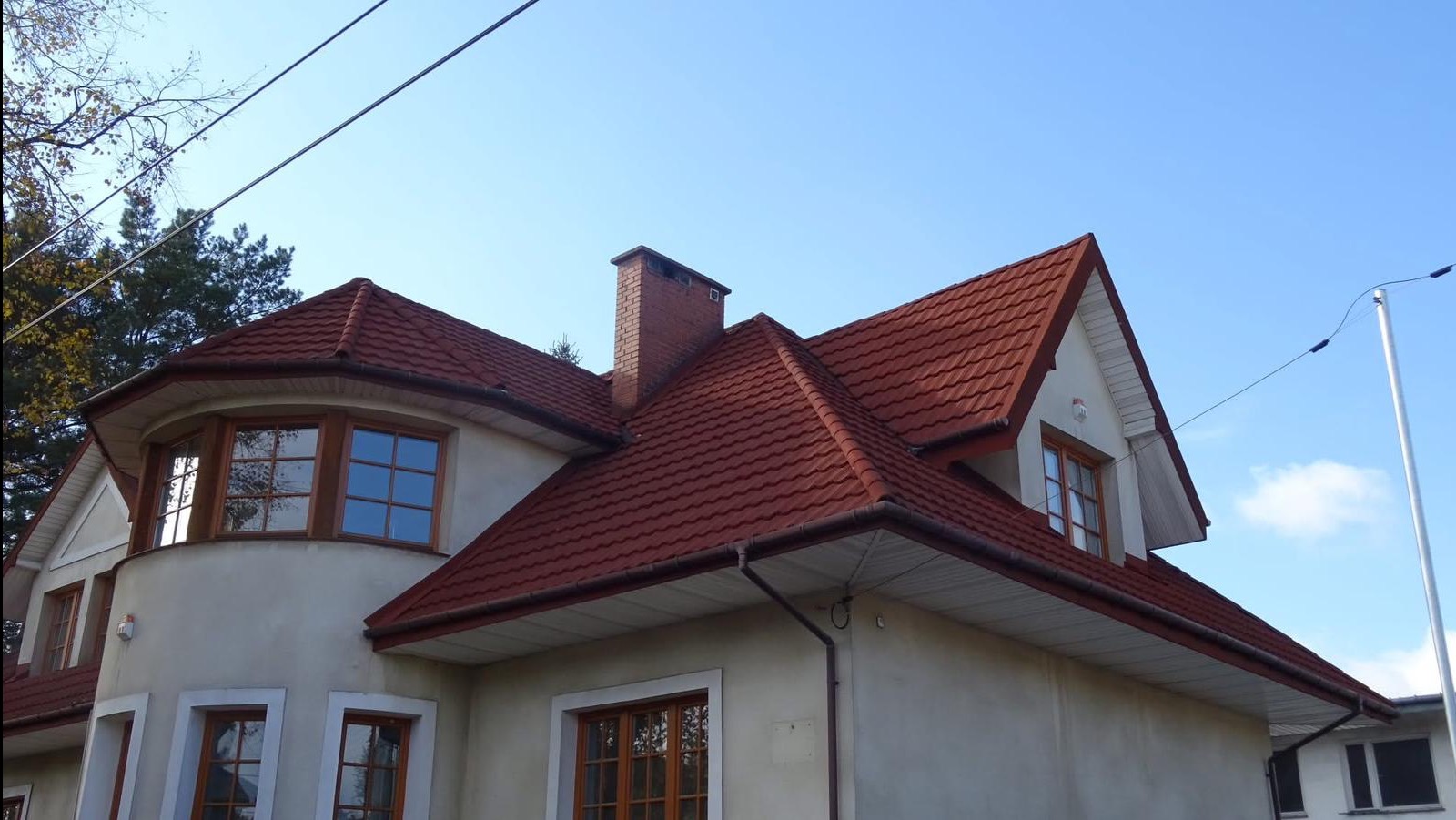 Dom z czerwoną dachówką i ceglanym kominem na tle błękitnego nieba. Widoczne okna z drewnianą stolarką i fragmenty elewacji. Ujęcie z dołu, podkreślające architekturę budynku.