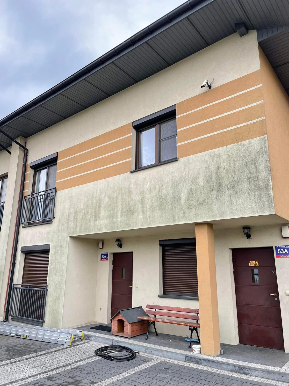 Dwukondygnacyjny budynek z elewacją w odcieniach beżu i brązu, z widocznymi śladami zabrudzeń na dolnej części. Na pierwszym planie drabina i wąż.