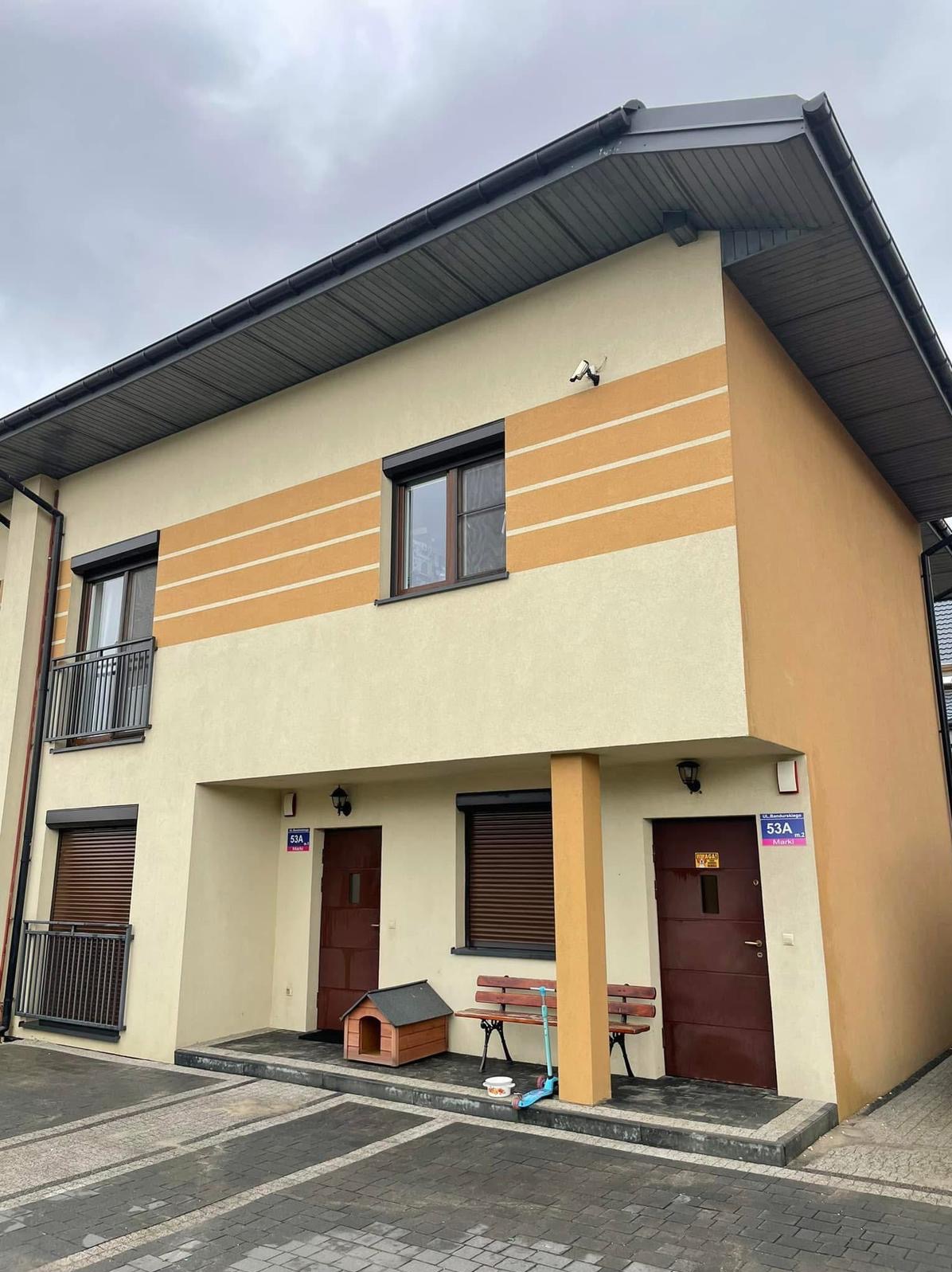 Dwukondygnacyjny budynek bliźniaczy z beżowo-brązową elewacją, brązowymi roletami i dachem. Widoczny fragment chodnika z kostki brukowej, ławka i buda dla psa.