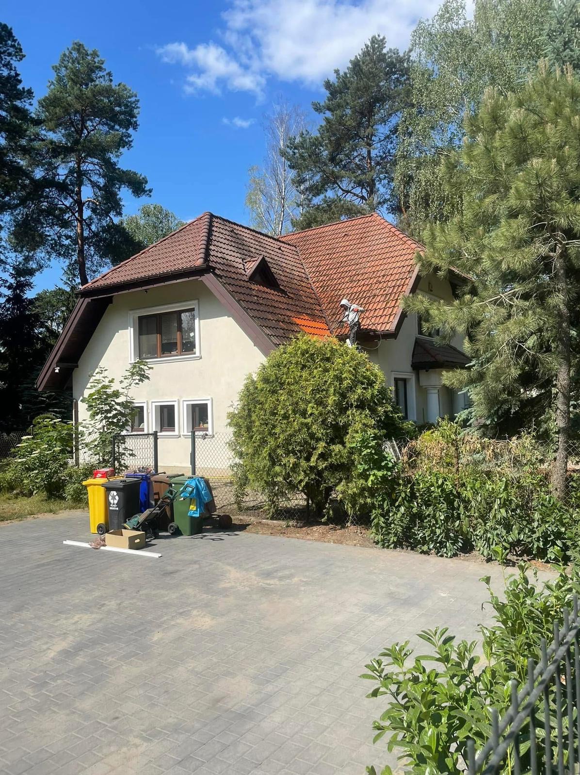 Pracownik w białym kombinezonie czyści dach domu z czerwonej dachówki za pomocą myjki ciśnieniowej. Widoczne zabrudzenia na dachu i otoczenie domu.