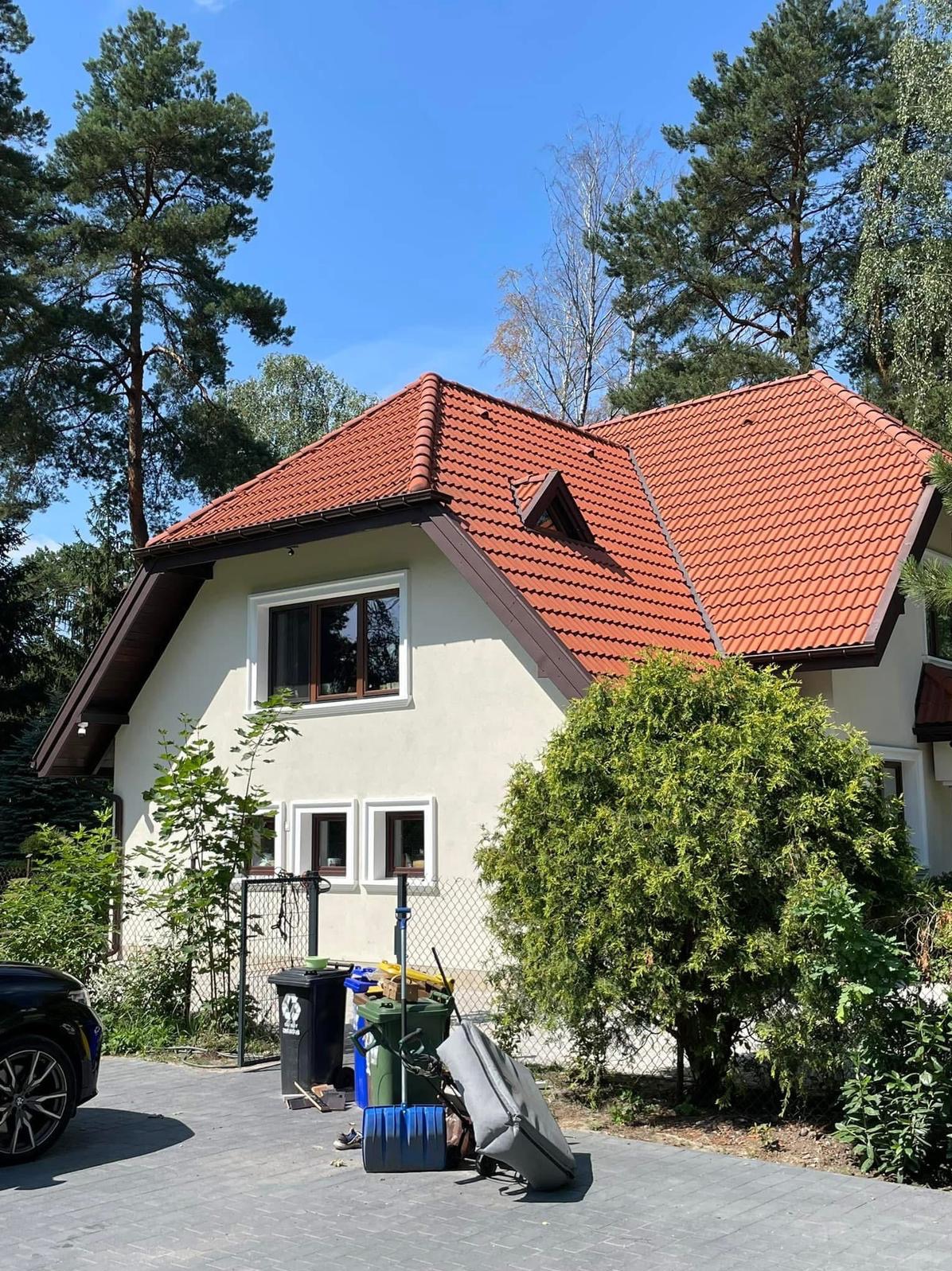 Dom jednorodzinny z czerwoną dachówką w Szczecinie, otoczony zielenią. Widoczne okna z brązowymi ramami i fragment podjazdu z koszami na śmieci.