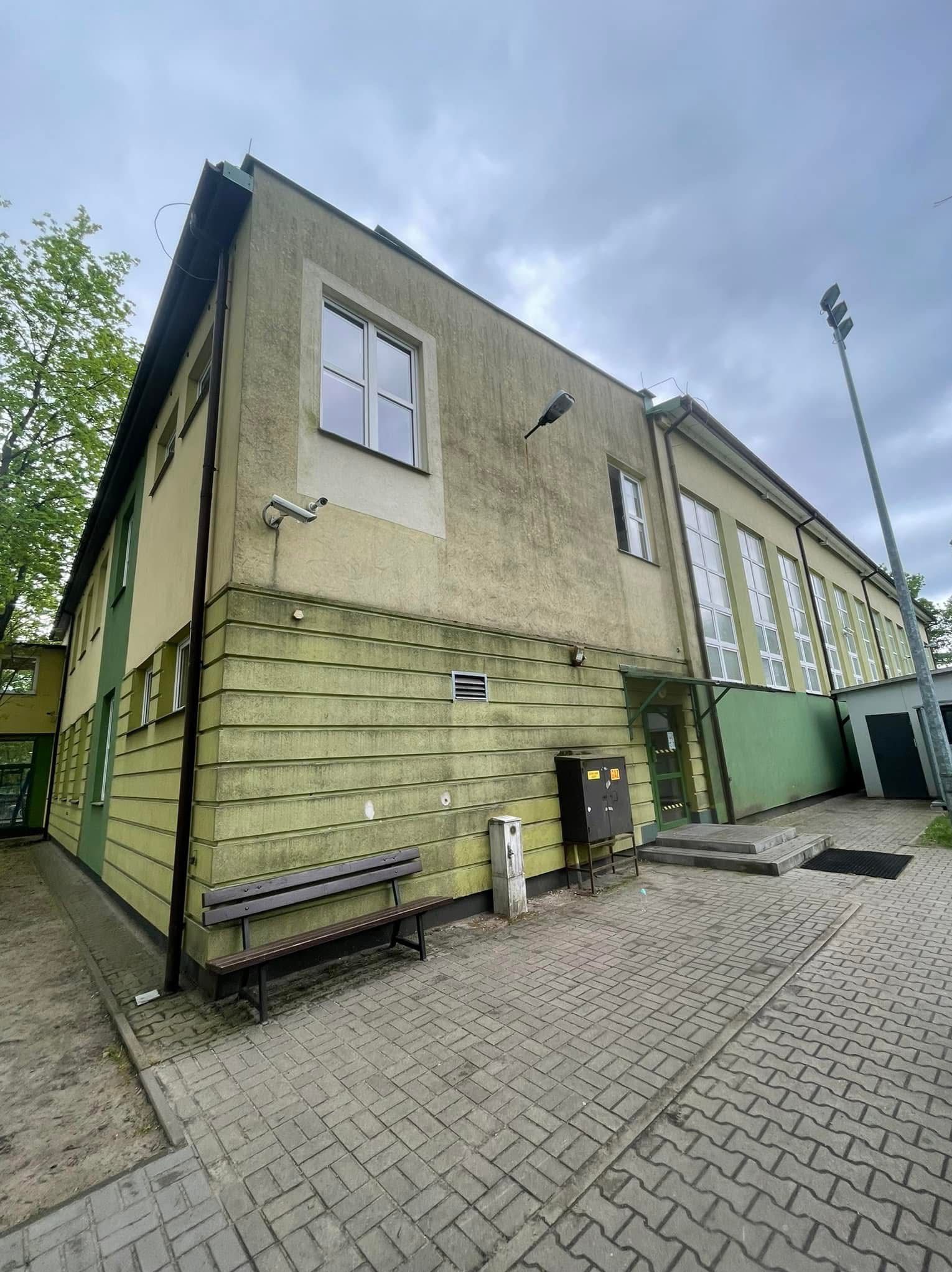 Dwukondygnacyjny budynek z elewacją w odcieniach zieleni i beżu, widoczna kamera, ławka i kostka brukowa. Budynek sprawia wrażenie wymagającego odświeżenia.