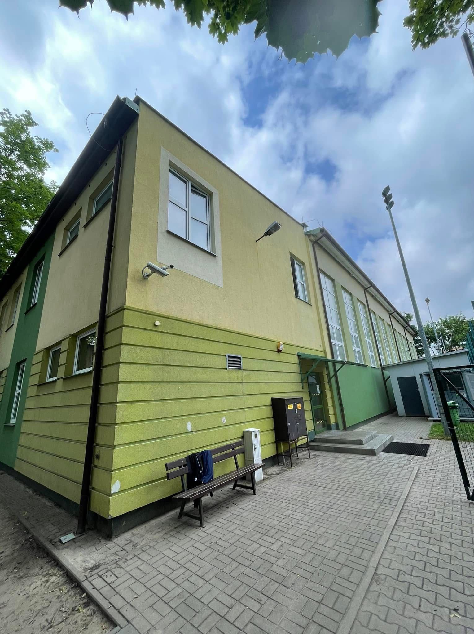 Dwukolorowa elewacja budynku z widoczną kamerą monitoringu, oświetleniem zewnętrznym i ławką z kurtką. Ujęcie z dołu, widoczne fragmenty nieba i drzew.