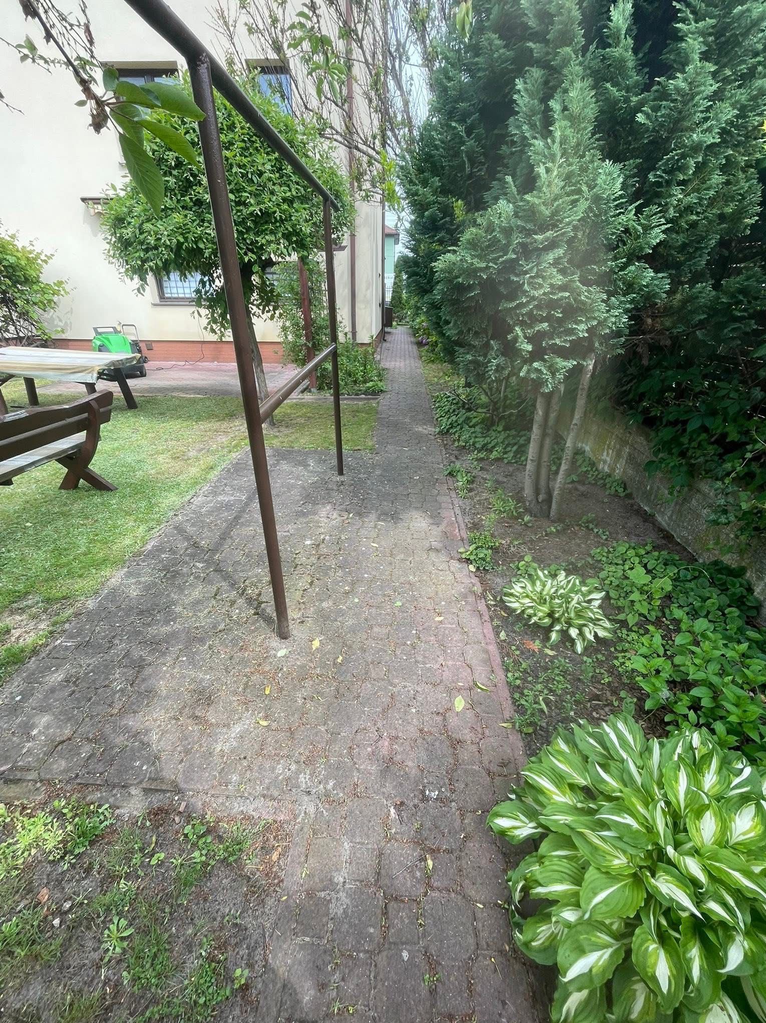 Zewnętrzna ścieżka z kostki brukowej, prowadząca przez ogród z zielenią i drzewami. Widoczne zabrudzenia na kostce, sugerujące potrzebę czyszczenia.