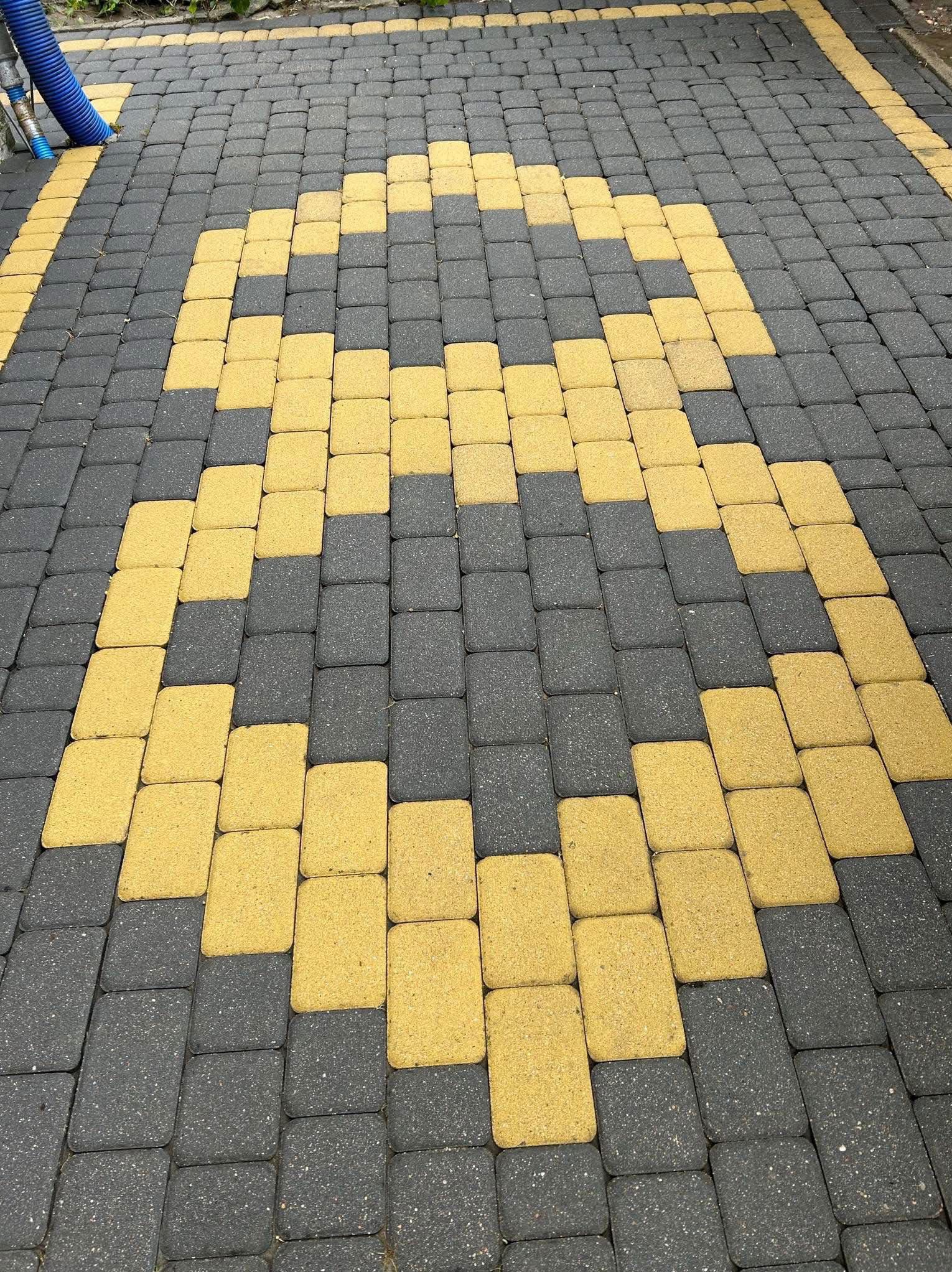 Ułożona kostka brukowa w odcieniach szarości i żółci tworząca wzór. W rogu widoczny fragment niebieskiego węża. Widok z góry, równomierne oświetlenie.