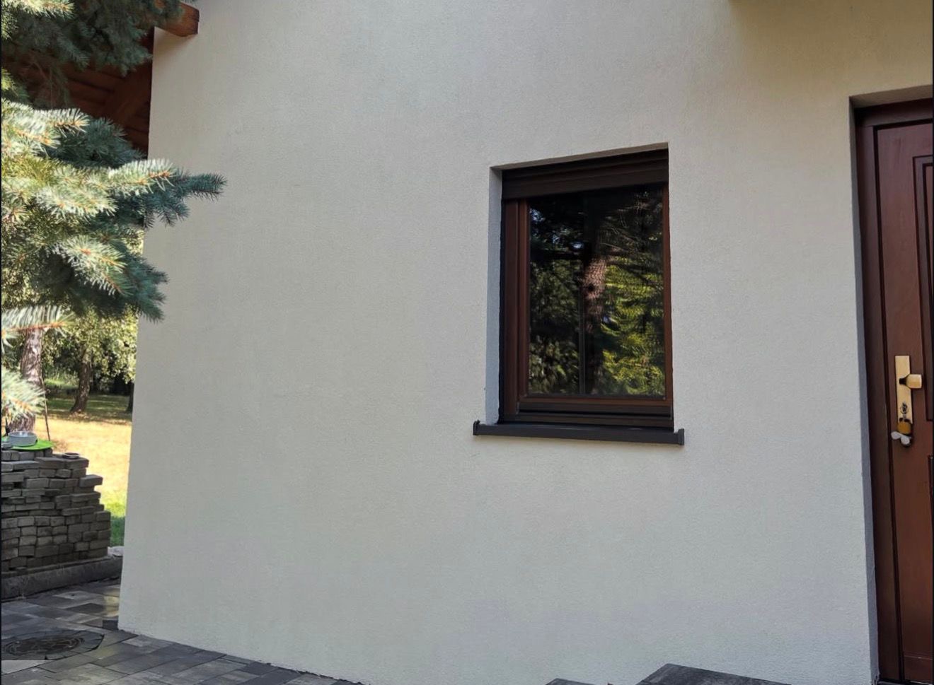 Jasna elewacja domu z brązowymi oknami i drzwiami. Widoczny fragment zielonego drzewa. Ujęcie z niskiej perspektywy, podkreślające fakturę ściany.