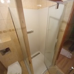 Fachura - Wykończona kabina prysznicowa z odpływem liniowym i sedesem z motywem muszli w łazience z beżowymi płytkami.