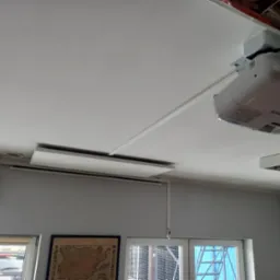Zamontowany projektor Epson na suficie z widocznymi korytkami instalacyjnymi maskującymi przewody, ekran projekcyjny zwinięty pod sufitem, klimatyzator na ścianie, widok z okna na samochody.