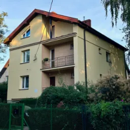 Dwupiętrowy dom po tynkowaniu elewacji z balkonem, widoczny zza zielonego ogrodzenia, z elementami dekoracyjnymi i świeżo pomalowaną fasadą w odcieniach beżu i brązu.