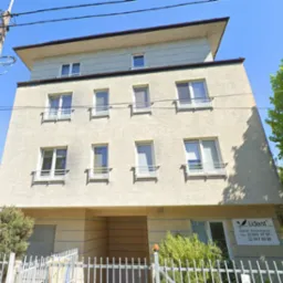 Dwupiętrowy budynek o beżowej, teksturowanej elewacji z białymi oknami i balustradami, widoczny z poziomu ulicy, z logo gabinetu stomatologicznego 'Lident' po prawej stronie.