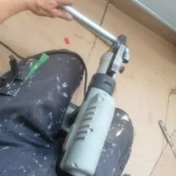 Hydraulik zaciska rurę metalową za pomocą elektrycznej zaciskarki, spodnie robocze poplamione farbą, kartonowe podłoże.