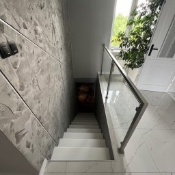 SG - BUD - Nowoczesne schody z gładzią dekoracyjną na ścianie, szklaną balustradą i białymi stopniami. Widok z góry na dół, z oknem i rośliną w tle.