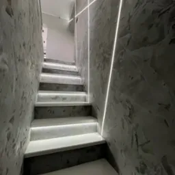 Schody z oświetleniem LED wzdłuż stopni i na ścianie o szarej, teksturowanej powierzchni. Nowoczesny design, minimalistyczny styl, chłodna kolorystyka.