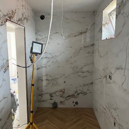 SG - BUD - Wykończona łazienka z marmurowymi płytkami i parkietem ułożonym w jodełkę. Widoczna lampa robocza na trójnogu. Częściowo widoczne przewody elektryczne.