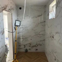 Wykończona łazienka z marmurowymi płytkami i parkietem ułożonym w jodełkę. Widoczna lampa robocza na trójnogu. Częściowo widoczne przewody elektryczne.