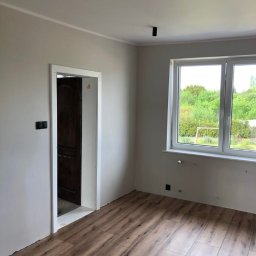 SG - BUD - Jasne wnętrze po remoncie: drewnopodobna podłoga, świeżo pomalowane ściany w odcieniach szarości i białe okno z widokiem na zieleń.