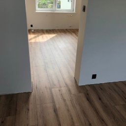 SG - BUD - Ukończone układanie paneli podłogowych w jasnym wnętrzu z oknem. Widoczne świeżo pomalowane ściany i gniazdka elektryczne. Perspektywa z progu pokoju.