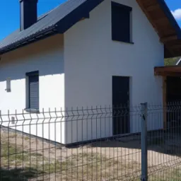 Nowoczesny dom jednorodzinny z białą elewacją, grafitowym dachem i roletami zewnętrznymi, widziany zza metalowego ogrodzenia panelowego.