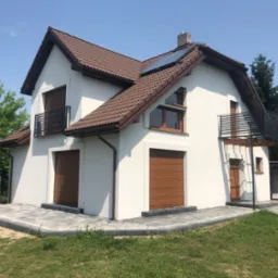 Nowoczesny dom jednorodzinny z białymi ścianami, brązową dachówką, roletami zewnętrznymi i balkonami z czarną balustradą, otoczony zielonym trawnikiem i drzewami.