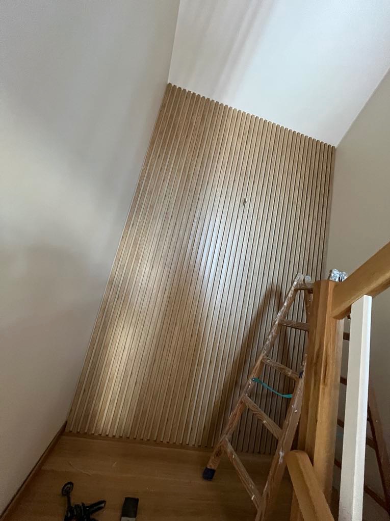 Drewniana boazeria lamelowa na ścianie klatki schodowej, widoczna drabina i narzędzia, co sugeruje trwający montaż. Naturalne oświetlenie podkreśla strukturę drewna.