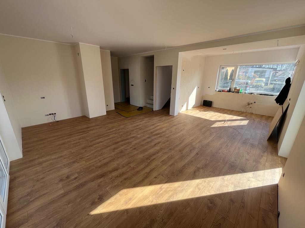 Wykończone drewniane panele podłogowe w przestronnym, jasnym pokoju podczas remontu. Widoczne przygotowane ściany do malowania i fragment okna z widokiem na zewnątrz.