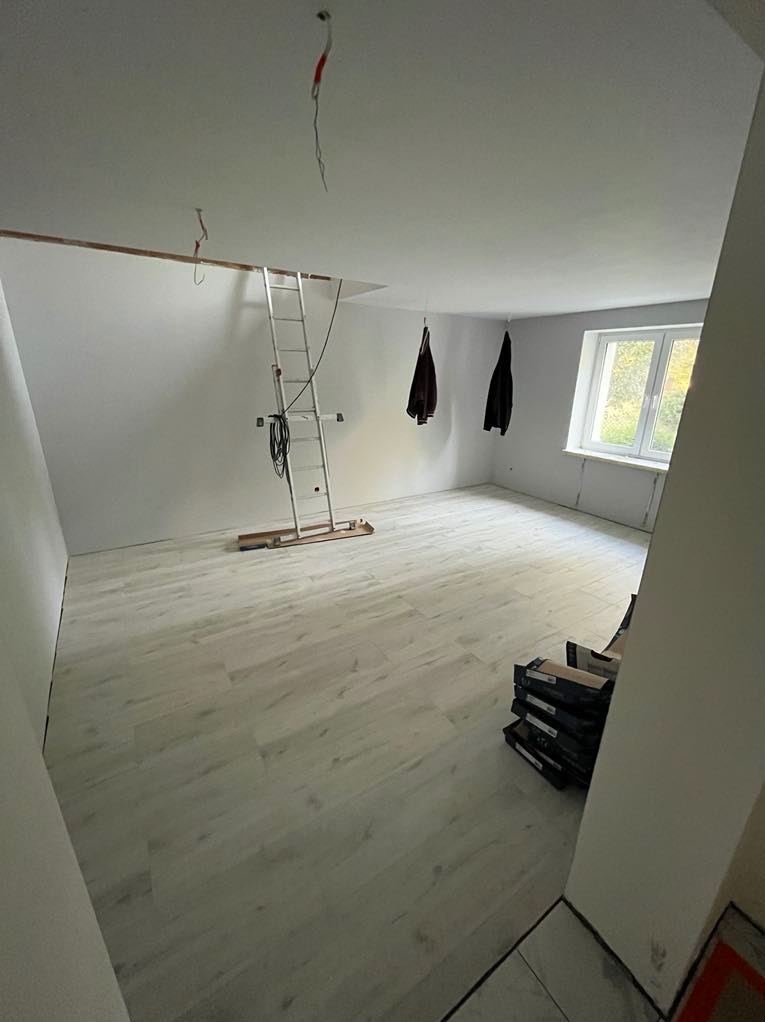 Jasne panele podłogowe w pokoju po remoncie, drabina oparta o ścianę, widoczne kable i wiszące ubrania. Okno z naturalnym światłem.