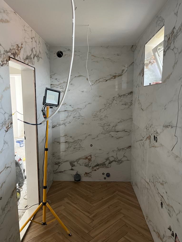 Wykończona łazienka z marmurowymi płytkami i parkietem ułożonym w jodełkę. Widoczna lampa robocza na trójnogu. Częściowo widoczne przewody elektryczne.