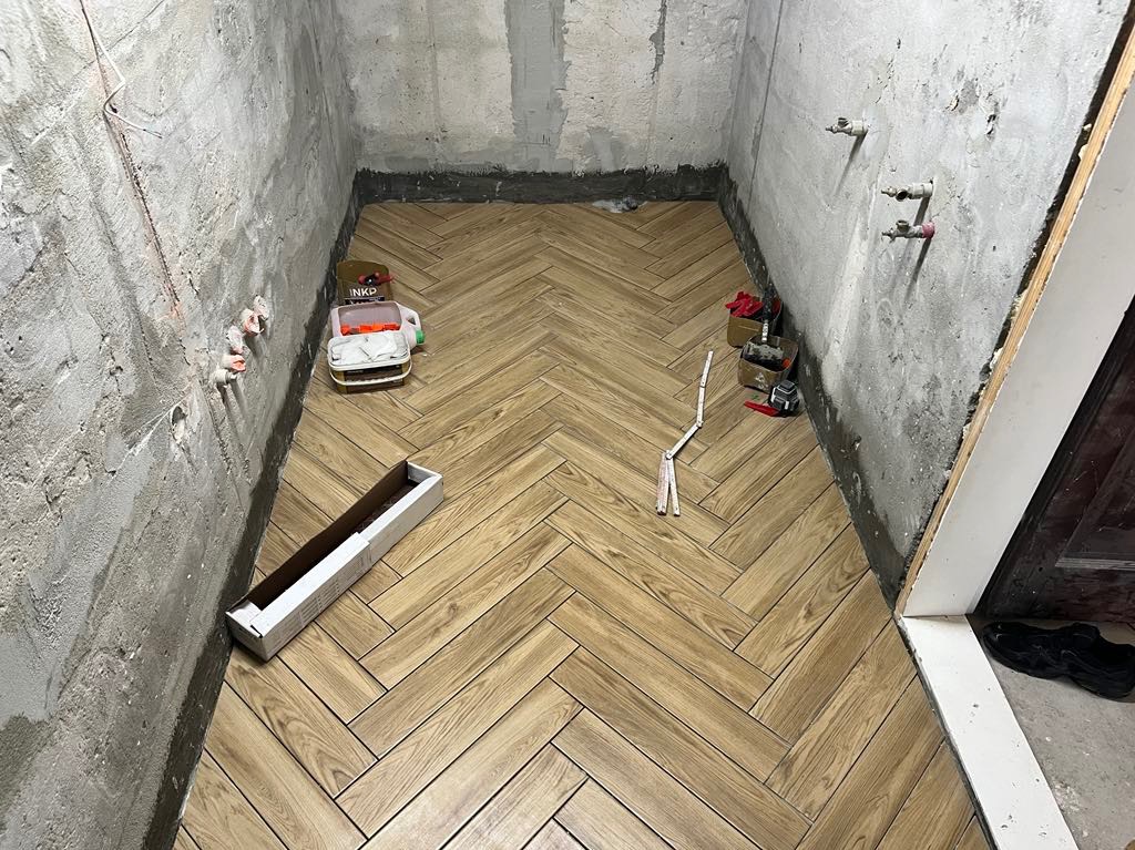 Ułożona drewnopodobna podłoga w jodełkę w surowej łazience. Widoczne narzędzia i materiały budowlane. Ściany w stanie deweloperskim.