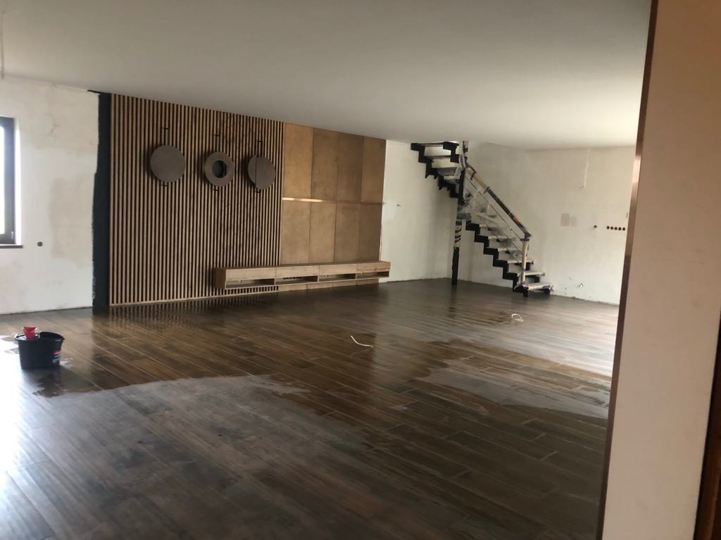 Salon po remoncie: parkiet w odcieniach brązu, dekoracyjna ściana z drewnianymi lamelami i okrągłymi ozdobami. Schody w tle, częściowo osłonięte folią.