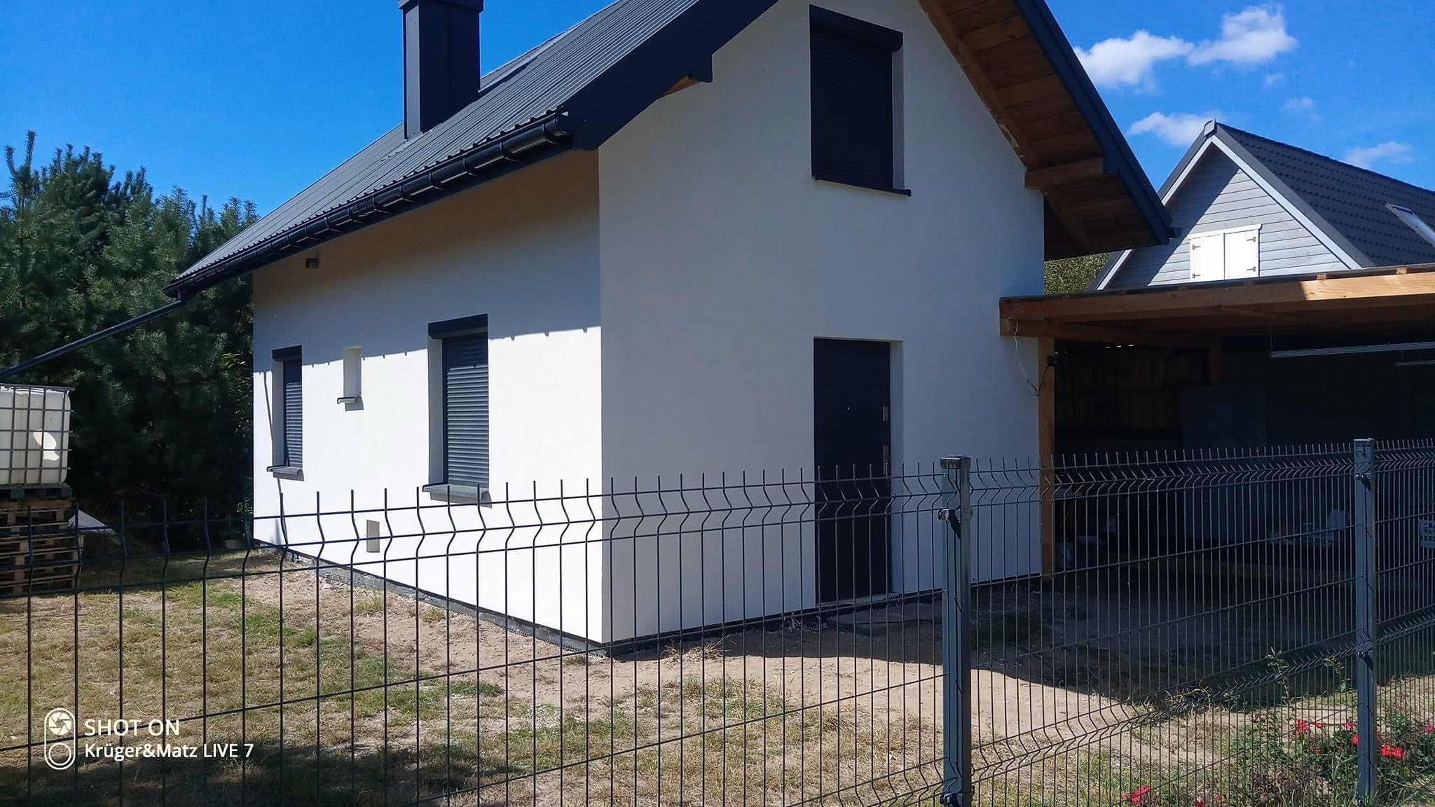 Nowoczesny dom jednorodzinny z białą elewacją, grafitowym dachem i roletami zewnętrznymi, widziany zza metalowego ogrodzenia panelowego.