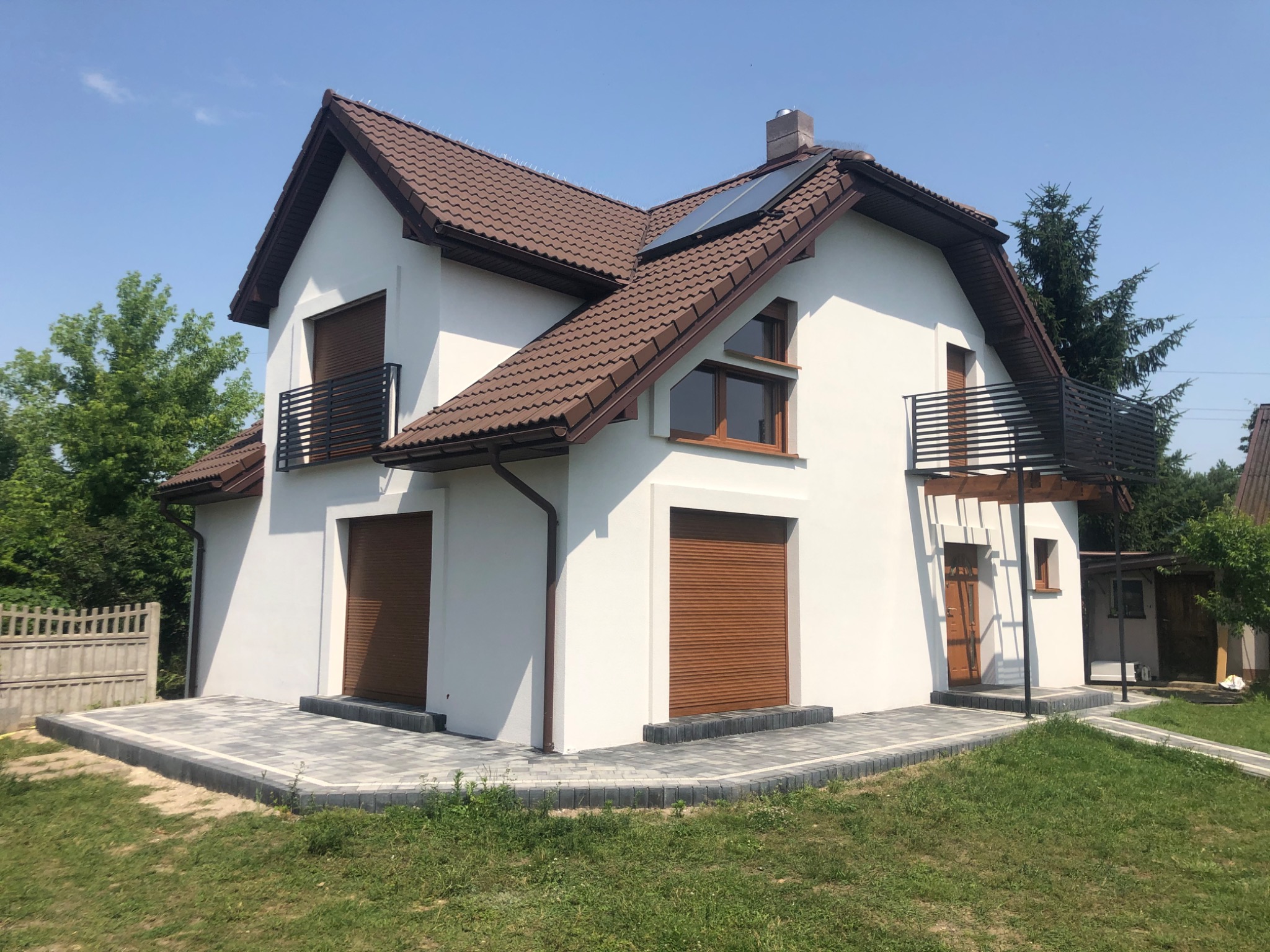 Nowoczesny dom jednorodzinny z białymi ścianami, brązową dachówką, roletami zewnętrznymi i balkonami z czarną balustradą, otoczony zielonym trawnikiem i drzewami.
