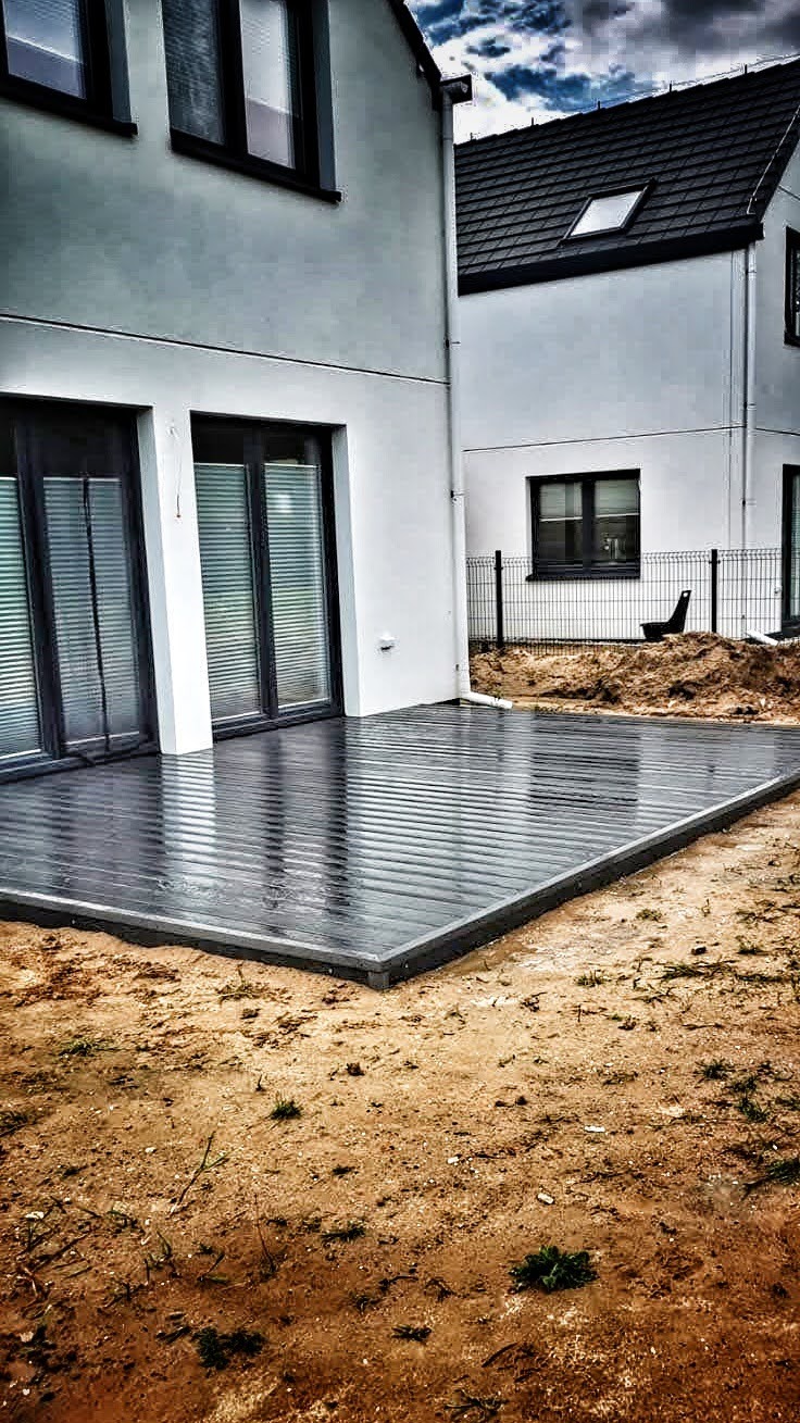 Nowoczesny taras z desek kompozytowych w kolorze grafitowym, przylegający do jasnego budynku z dużymi oknami, widoczny fragment ogrodu z ziemią.