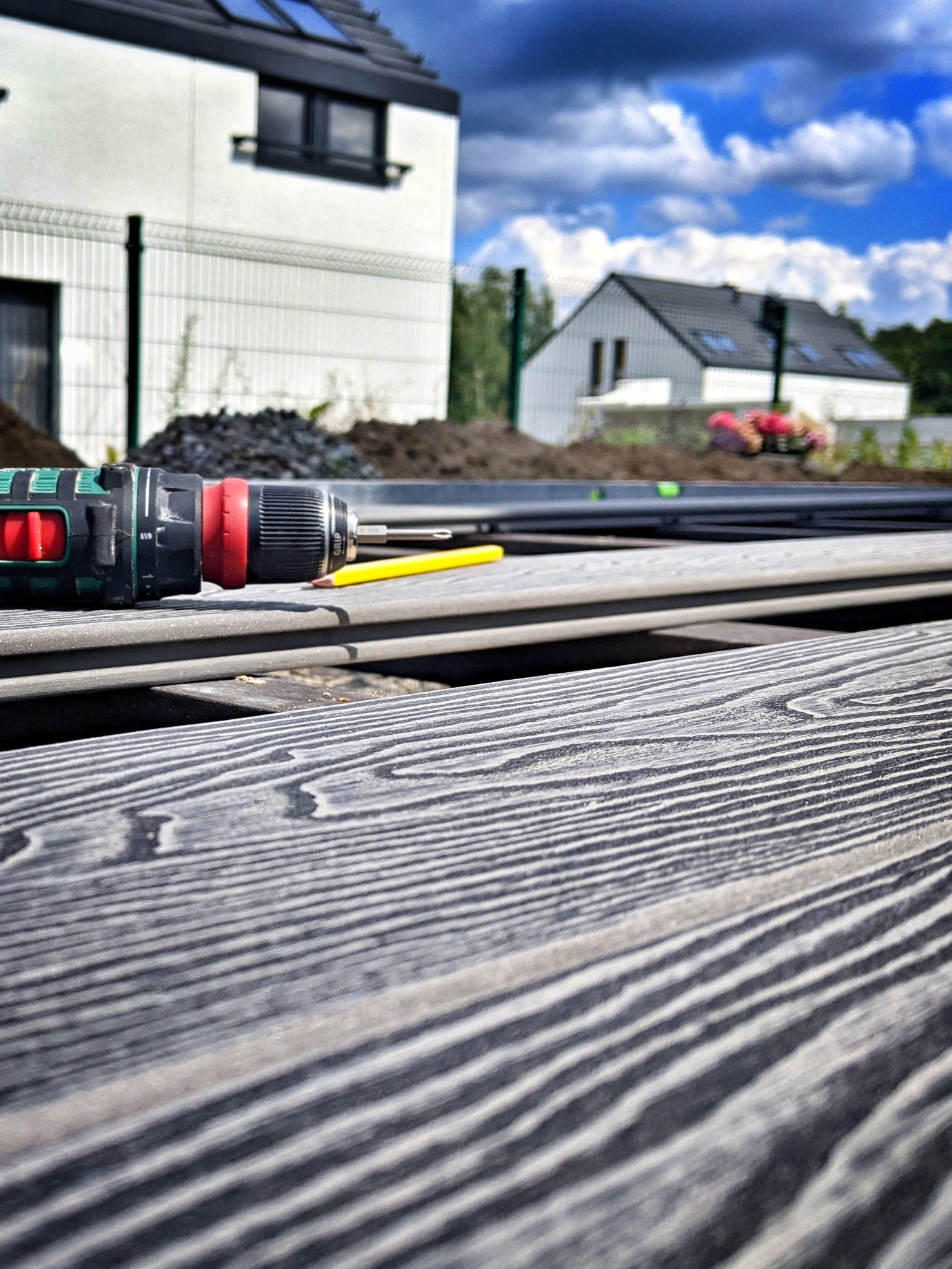 Montaż desek kompozytowych na tarasie: zbliżenie na wkrętarkę, ołówek i deski z widoczną strukturą drewna, w tle dom jednorodzinny i przygotowany teren pod ogród.