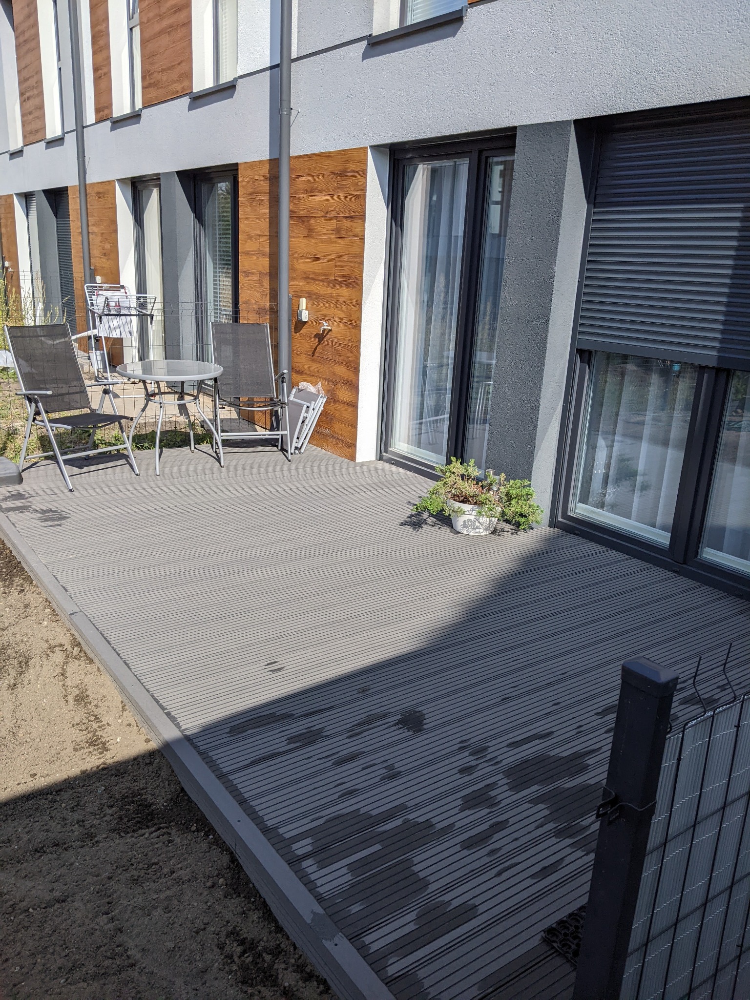 Szary taras kompozytowy przy nowoczesnym domu z szarą elewacją i drewnianymi akcentami, z meblami ogrodowymi i doniczką z zielenią, widok z niskiej perspektywy.