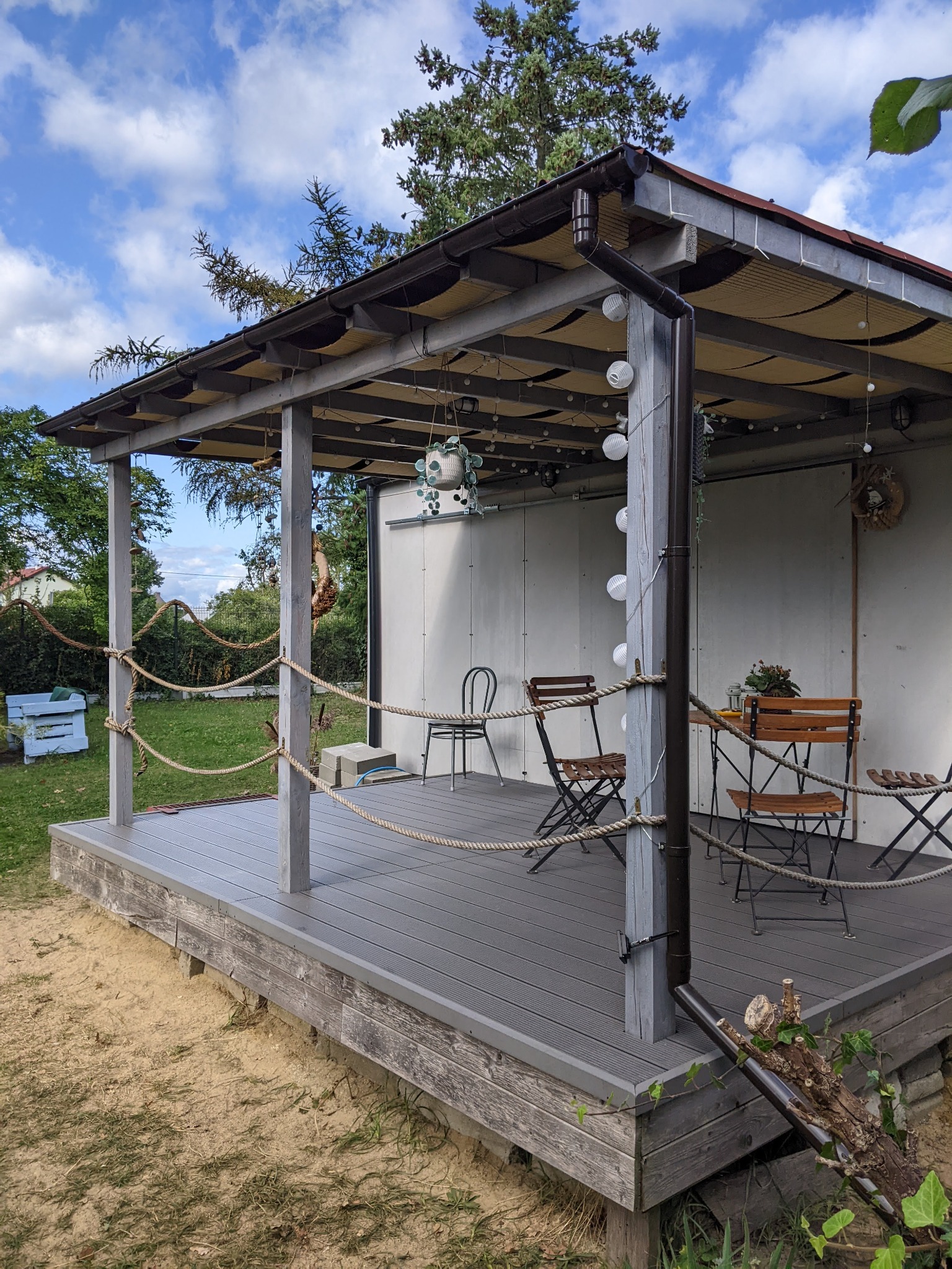 Zadaszony taras kompozytowy z drewnianymi słupami, obwiązany liną jutową jako balustradą, w tle widoczny trawnik i zabudowania gospodarcze.