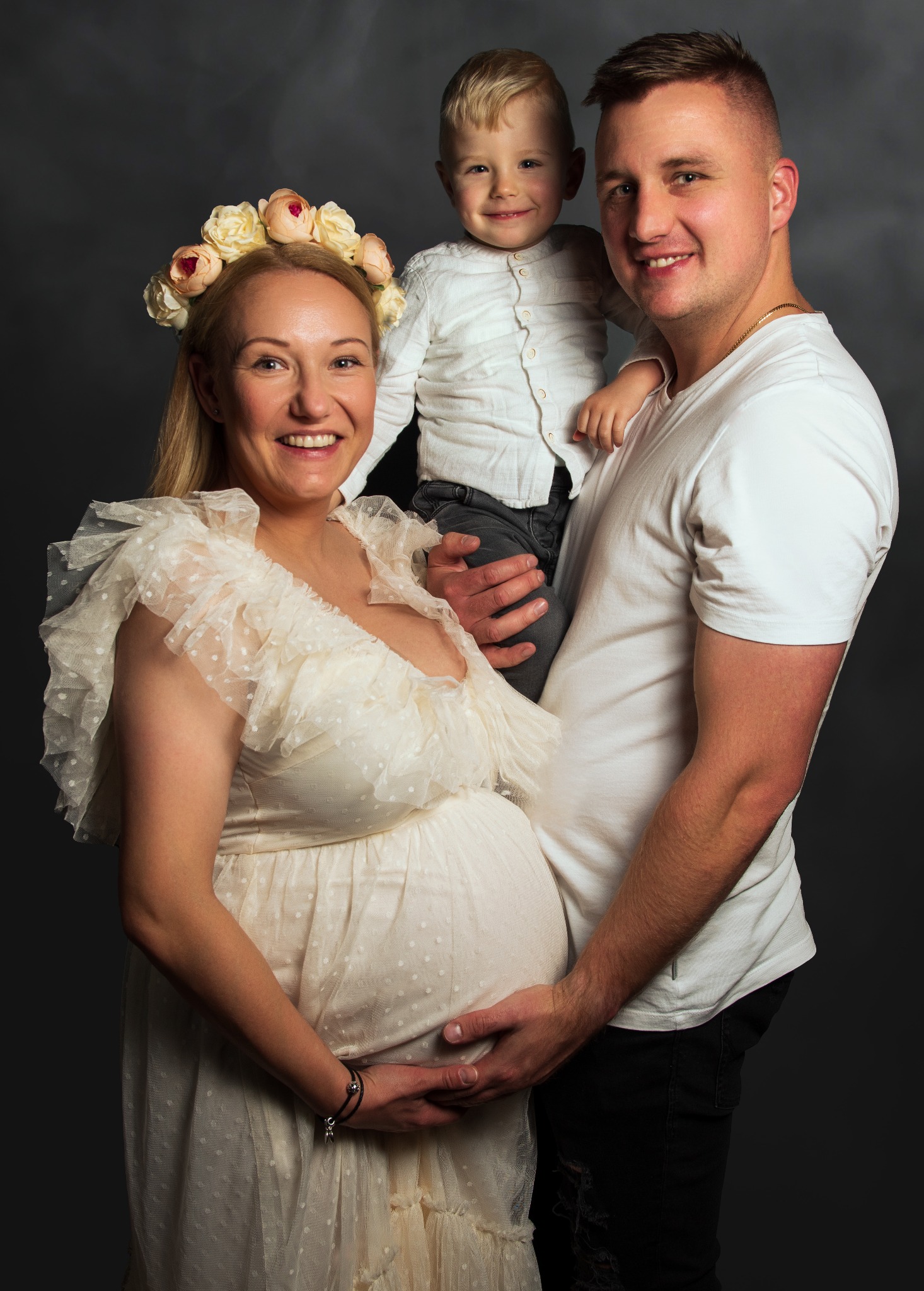 Uśmiechnięta, ciężarna kobieta w kremowej sukience z falbankami i wiankiem z róż na głowie, pozuje z mężem trzymającym synka na rękach na ciemnoszarym tle.