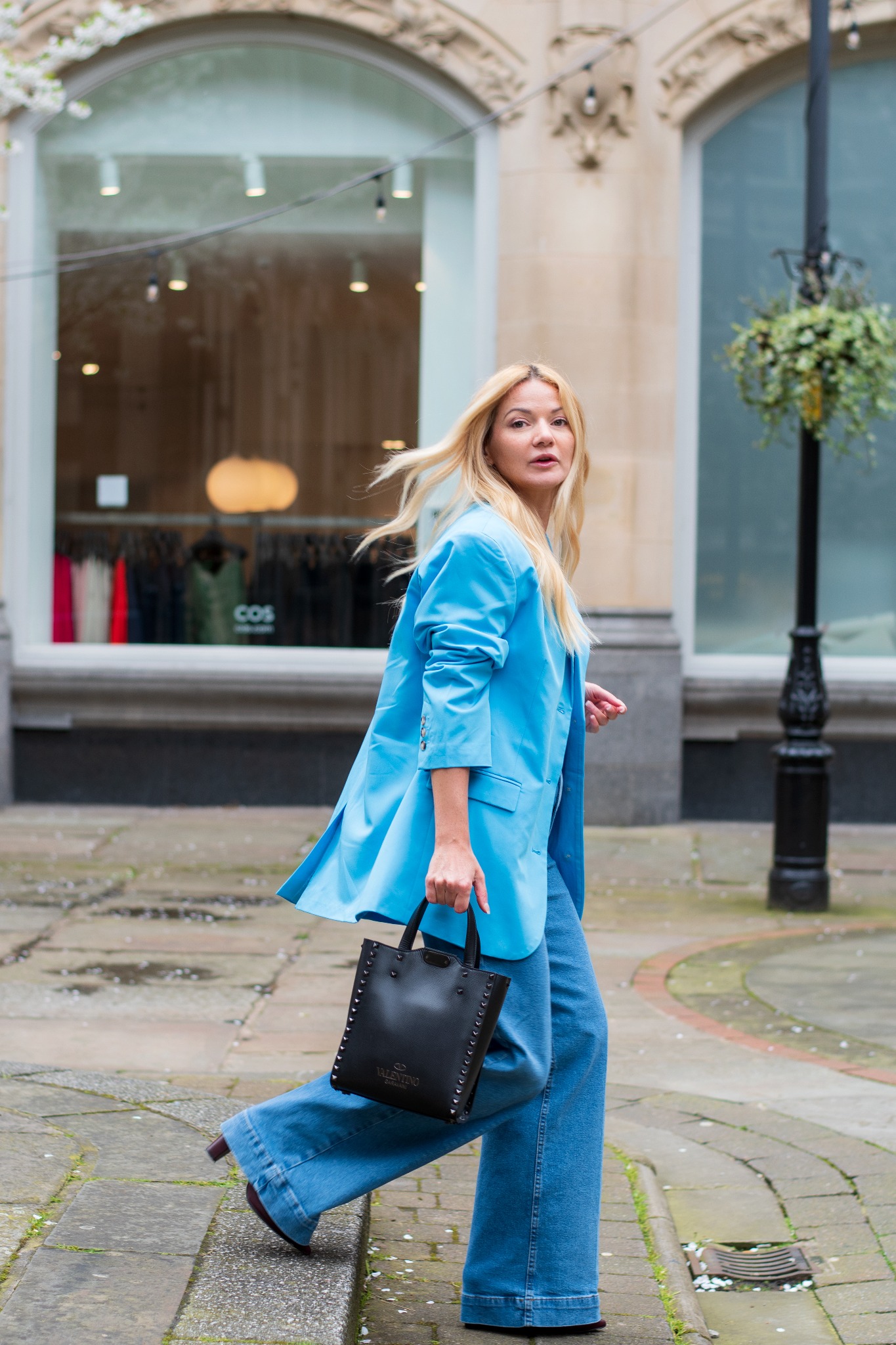 Kobieta z rozwianymi blond włosami w błękitnej marynarce i szerokich jeansach idzie po chodniku, trzymając czarną torebkę z ćwiekami na tle witryny sklepowej.
