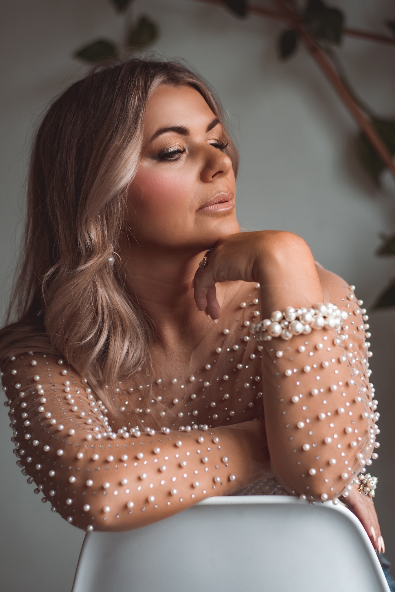 Portret kobiety z blond włosami, w bluzce ozdobionej perłami, opierającej podbródek na ręce z perłową bransoletką i pierścionkiem, patrzącej w dół.