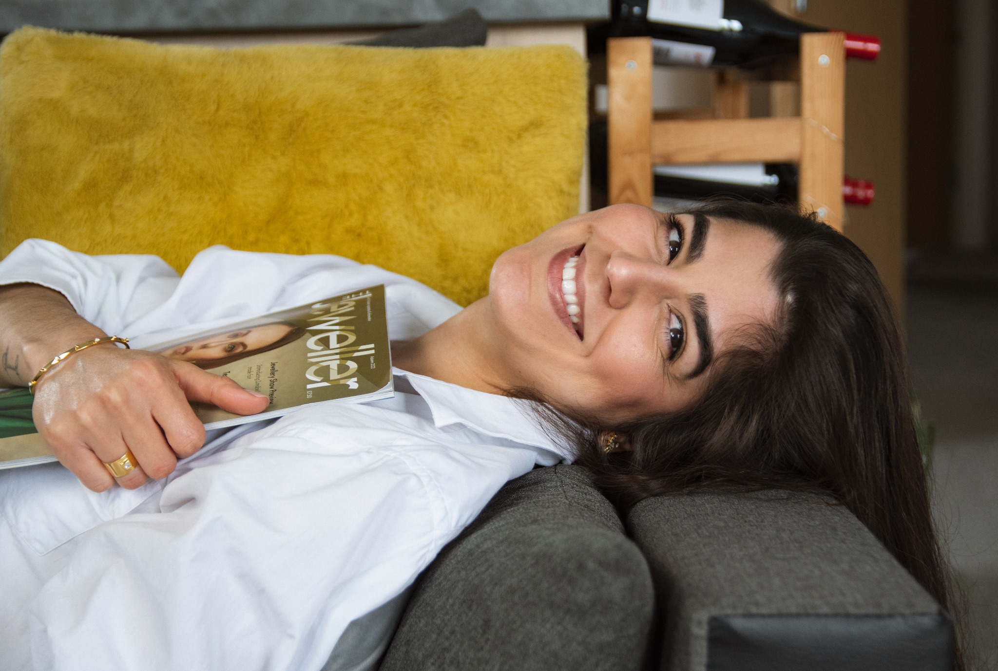 Uśmiechnięta kobieta w białej koszuli leży na szarej sofie z żółtą poduszką, trzymając magazyn o tematyce jubilerskiej i prezentując biżuterię na dłoni.
