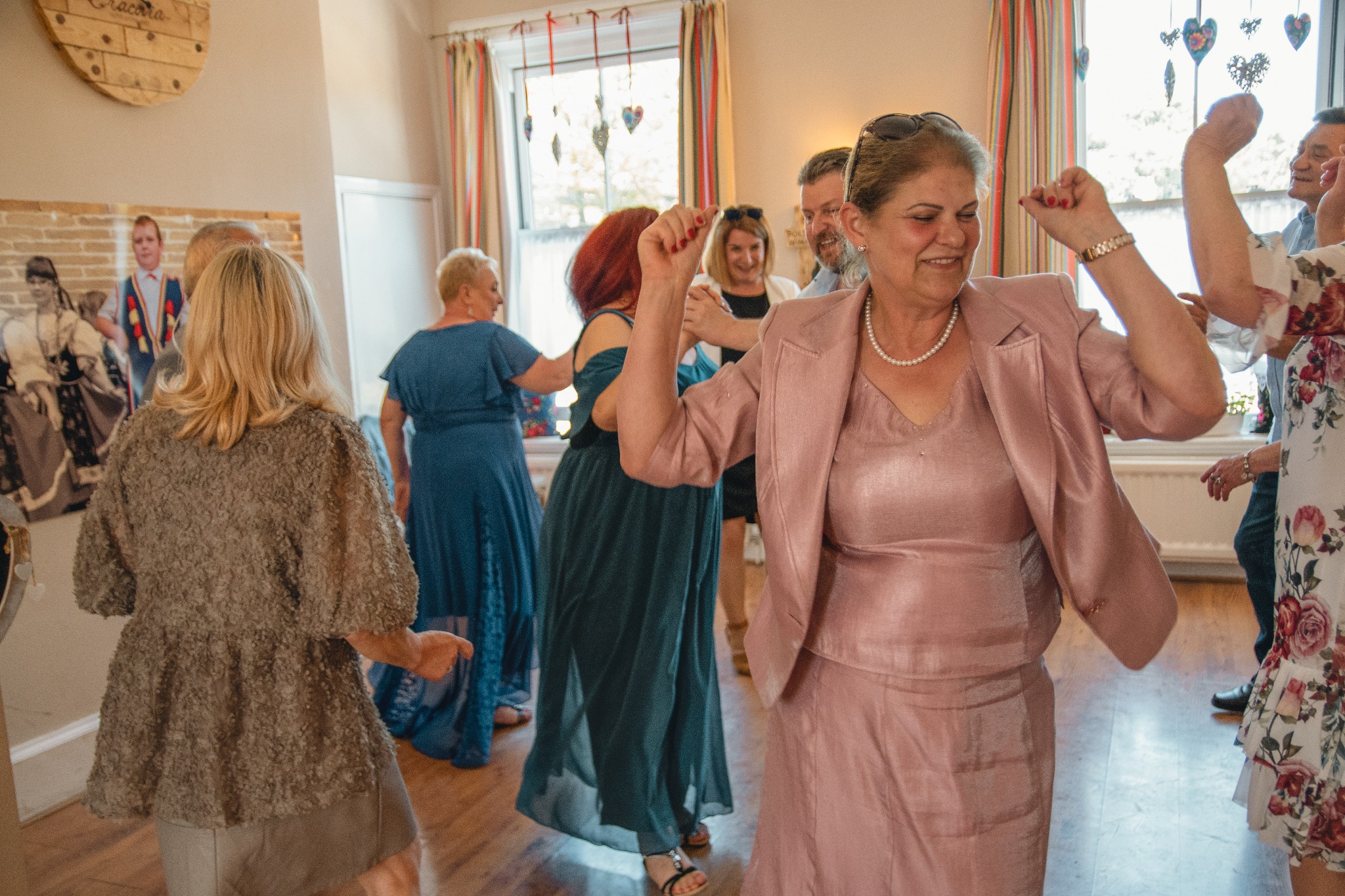 Grupa osób tańczy w jasnym pomieszczeniu; kobieta w różowym kostiumie na pierwszym planie, uśmiechnięta, z podniesionymi rękami, w tle okno z dekoracjami w kształcie serc.