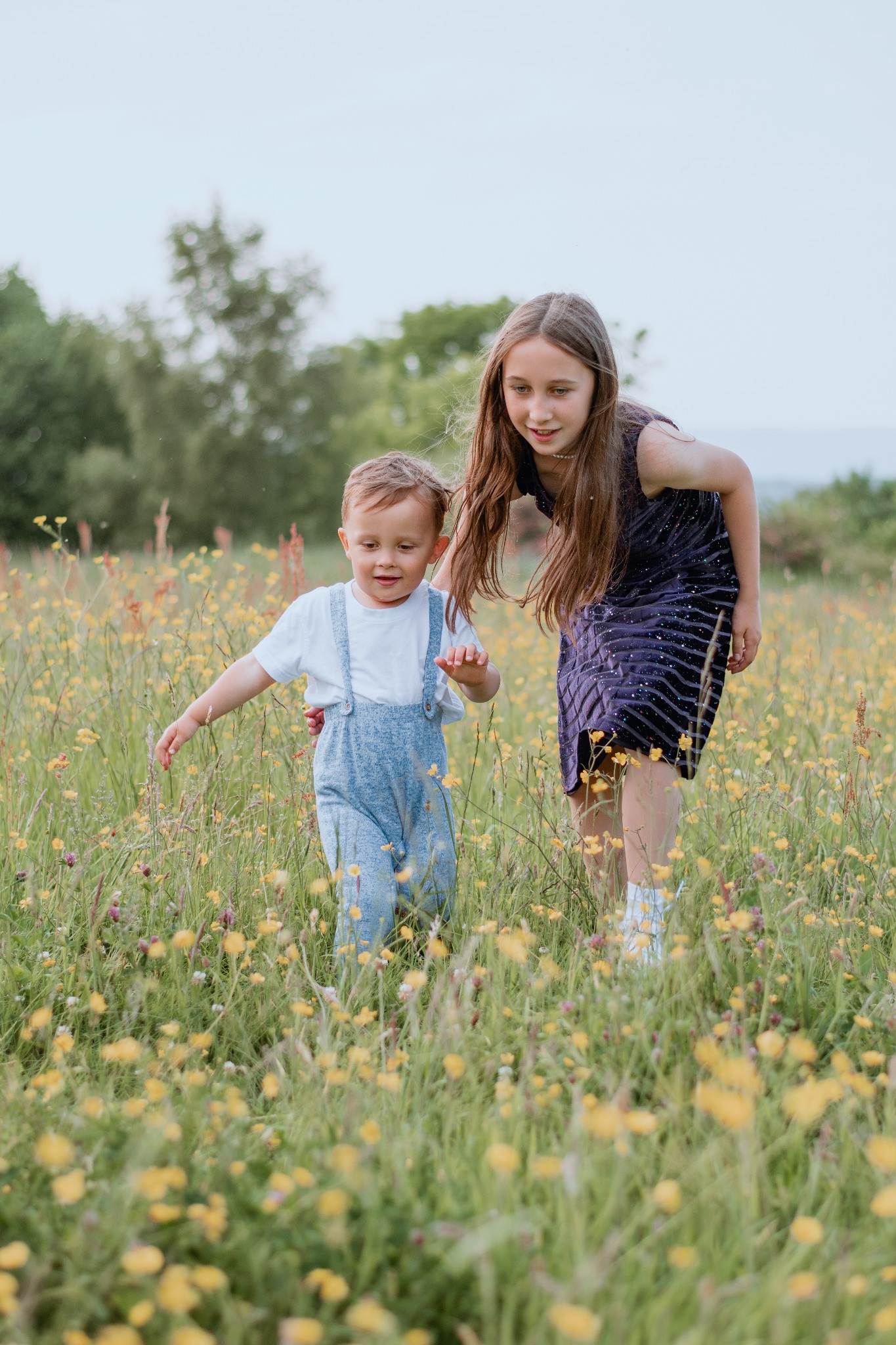 Chłopiec w ogrodniczkach prowadzony przez dziewczynkę w sukience po łące pełnej żółtych kwiatów.