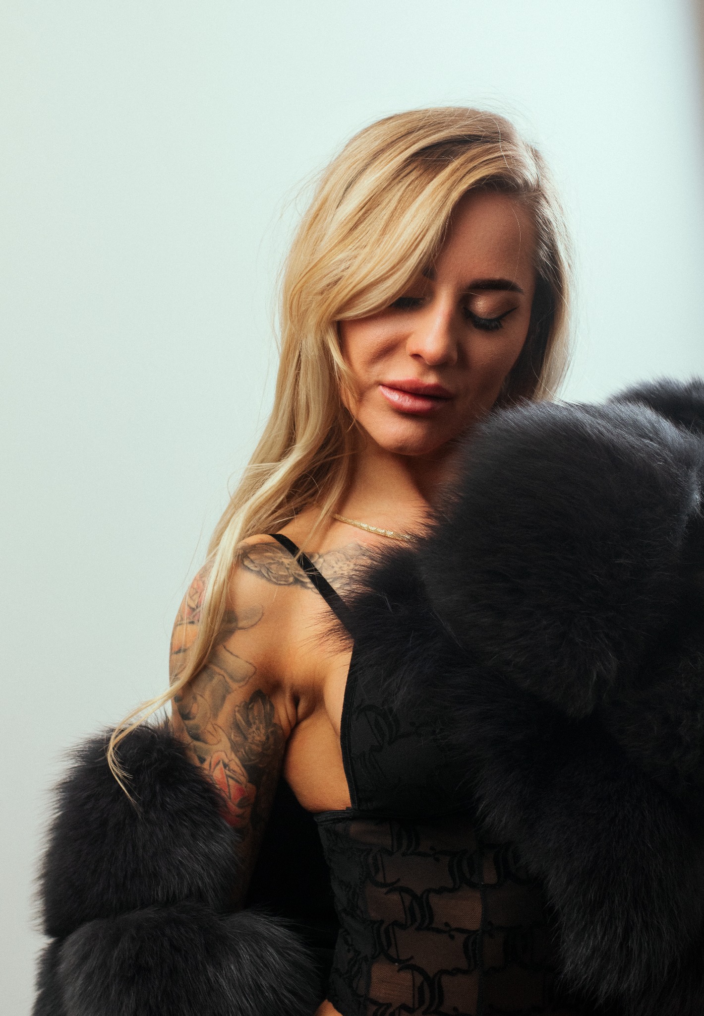 Portret kobiety z blond włosami, w czarnej bieliźnie i futrze, z tatuażami na ramieniu, spoglądającej w dół.
