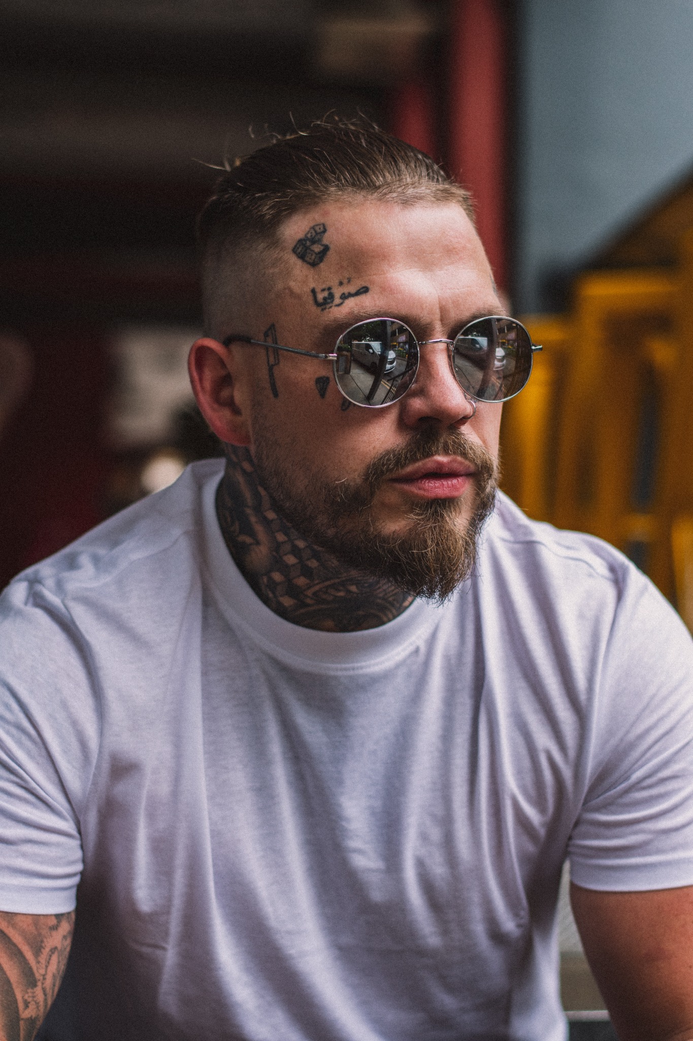 Portret mężczyzny z brodą i tatuażami na twarzy i szyi, w okularach przeciwsłonecznych odbijających ulicę, ubrany w białą koszulkę.