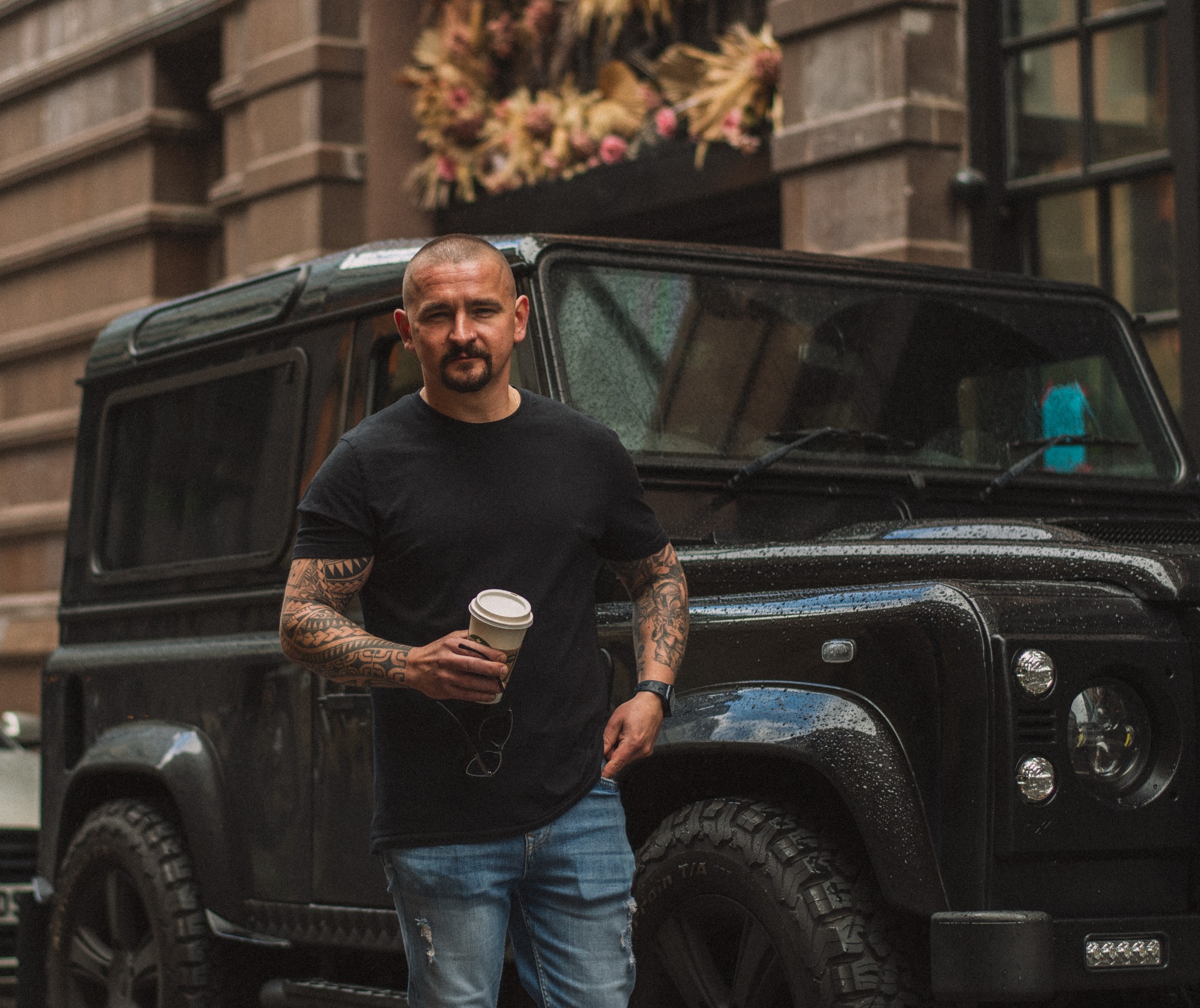 Mężczyzna z tatuażami na ramionach, trzymający kubek z kawą, stoi obok czarnego, terenowego samochodu na tle budynku z dekoracją kwiatową nad wejściem.