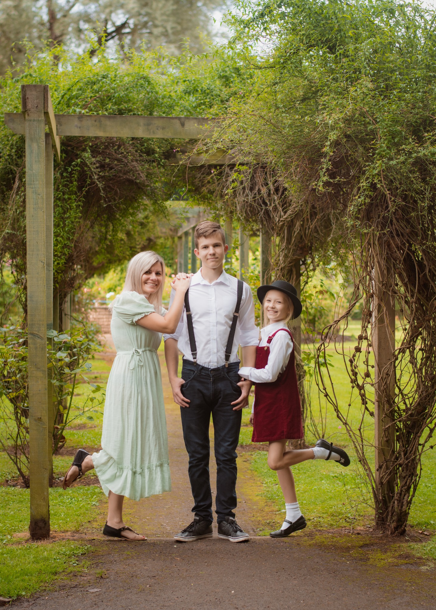 Uśmiechnięta rodzina: mama w zwiewnej sukience, nastolatek w szelkach i dziewczynka w sukience i kapeluszu, pozują na tle porośniętej pergoli w parku.