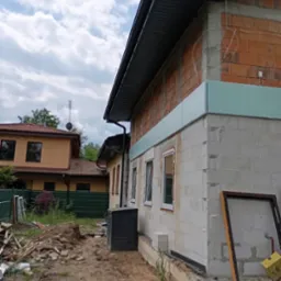 Budynek w trakcie ocieplania: widoczne bloczki, styropian, okna, rynna, oraz fragment dachu z ciemną dachówką, z elementami rozgardiaszu budowlanego w otoczeniu.