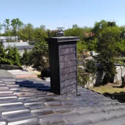 Ciemny, nowy komin z metalową czapą, wystający z błyszczącego, brązowego dachu ceramicznego w słoneczny dzień.