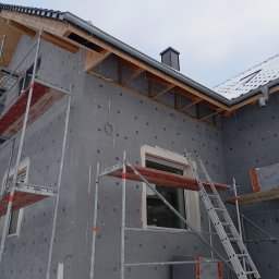 Oleksandr Vasiliev DACHOLEK - Dom w trakcie ocieplania: szare płyty styropianowe z charakterystycznymi kropkami, rusztowania, drabina oparta o ścianę, widoczne rynny i fragment dachu pokrytego śniegiem.